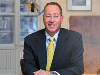 The image shows a man wearing glasses, a suit, and a tie, standing behind a desk with various items on it.