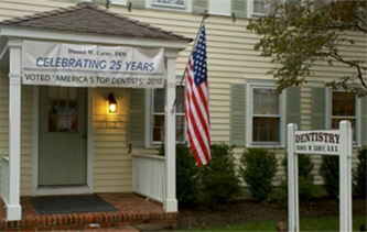 The image shows a small, white building with a sign indicating  Celebrating 25 Years  and a flag hanging from the roof. A banner on the building reads  Voted America s Top Dentist, 2013.  There is a sign in front of the building that says  DENTIST  and another sign with a phone number. The building has a porch with a door, and there are American flags displayed on the porch.