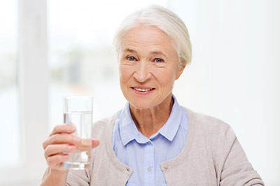 The image shows an elderly woman holding a glass of water with both hands, smiling at the camera.