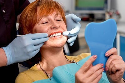 A woman with red hair sitting in a dental chair, smiling at the camera while holding up a blue mouthguard, with a dentist examining her teeth using a mirror and a probe.