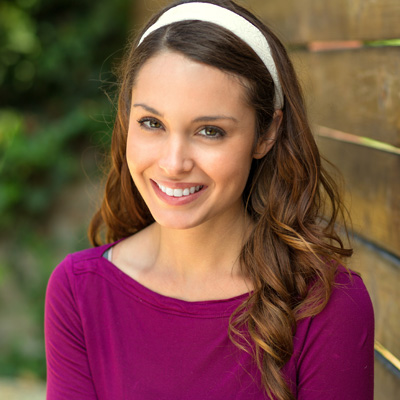 A smiling woman with long brown hair and a purple top poses against a wooden fence.