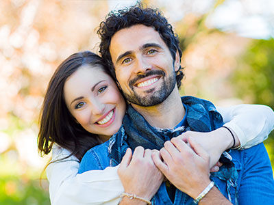 A man and woman are embracing each other outdoors with a tree in the background.