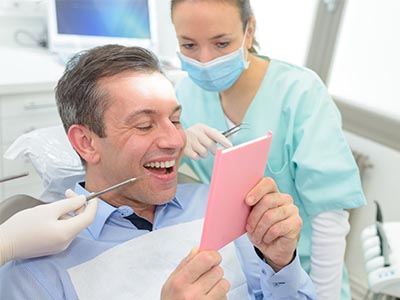 The image shows a man sitting in a dental chair with his eyes closed while holding a pink card, smiling broadly  he appears to be receiving dental care from a professional who is standing behind him, wearing a surgical mask and holding dental instruments.