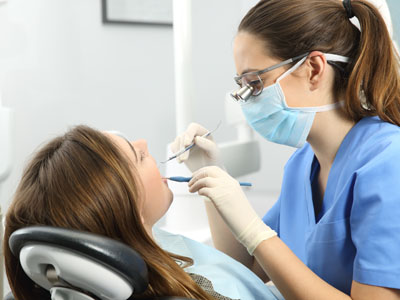 This image shows a dental hygienist performing a teeth cleaning procedure on a patient while wearing personal protective equipment  PPE , including gloves, a mask, and an apron.