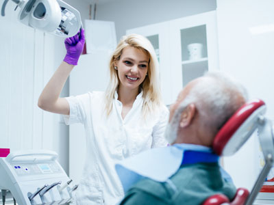 A dental hygienist in a white coat is assisting an elderly man with a chin strap during his dental appointment, with both wearing face masks.