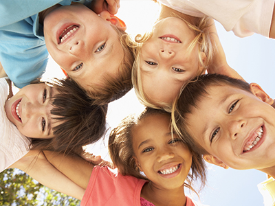 The image shows a group of children smiling at the camera while standing close together outdoors on a sunny day.