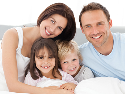 The image depicts a family of four, including two adults and two children, all smiling and posing together on a bed with a white sheet.