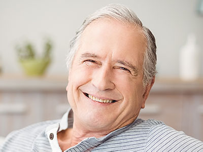 The image shows a smiling older man with grey hair, wearing a blue shirt, sitting comfortably in an indoor setting with a warm ambiance.