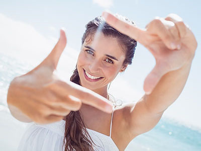 A woman with long hair smiles while holding her hand up to her face, creating a frame with her fingers.